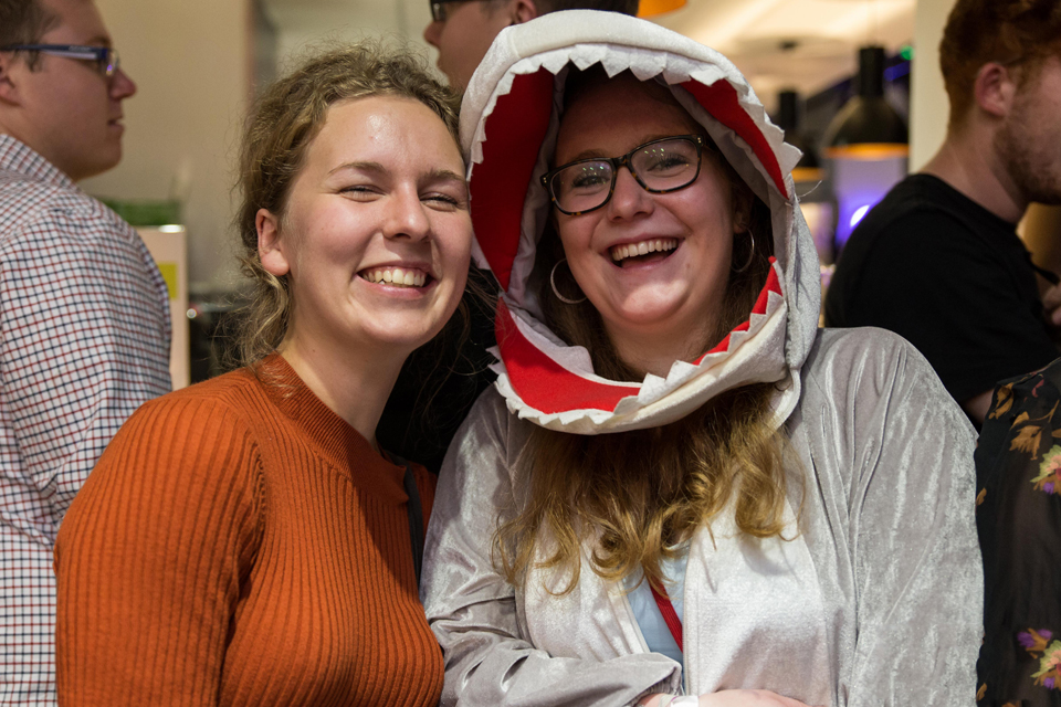 Two female students, one of them wearing a shark costume, smiling and laughing, with other students in the background.